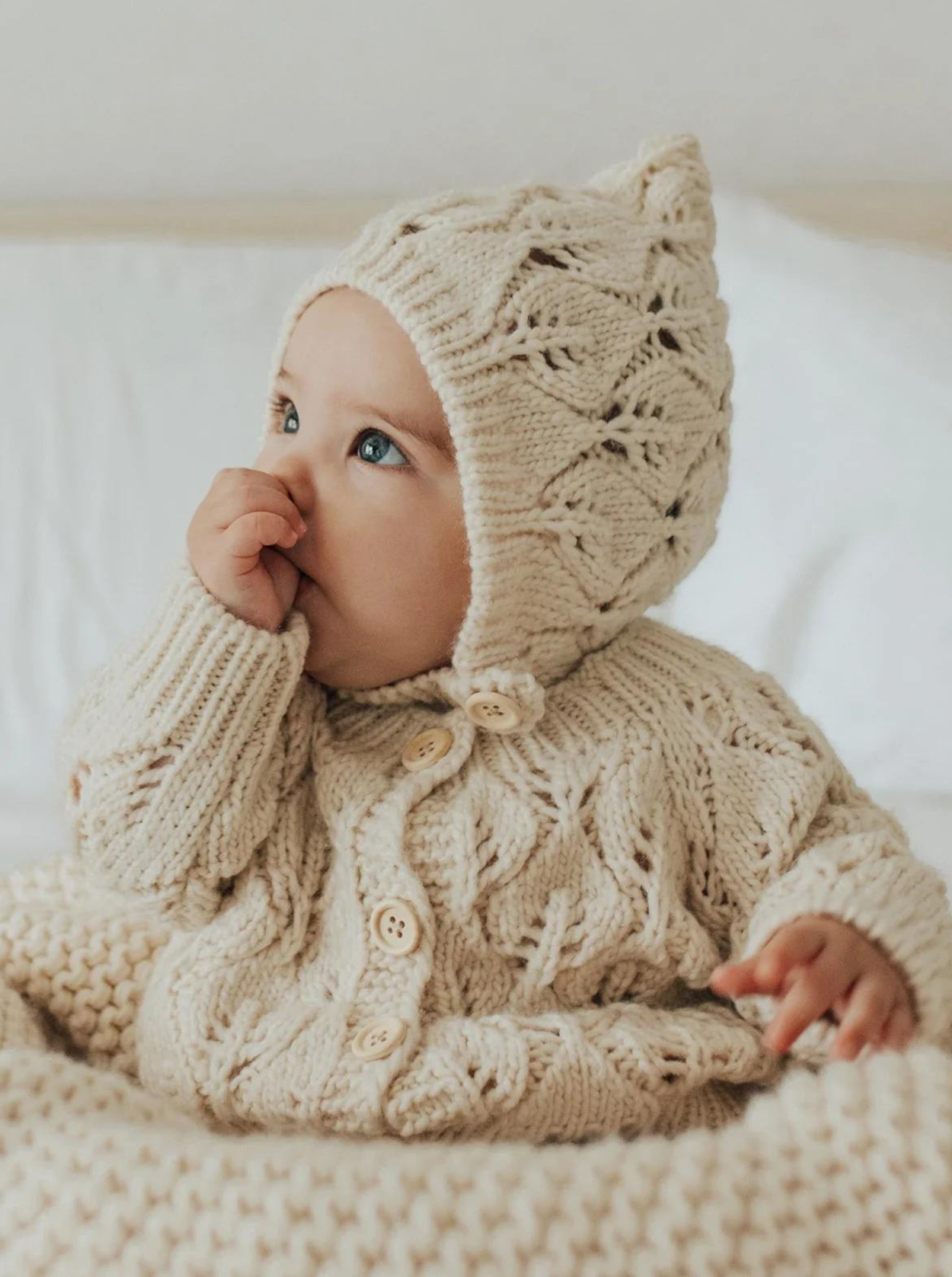 Baby in a beige knitted outfit and hat, sitting and sucking their thumb on a cozy blanket.
