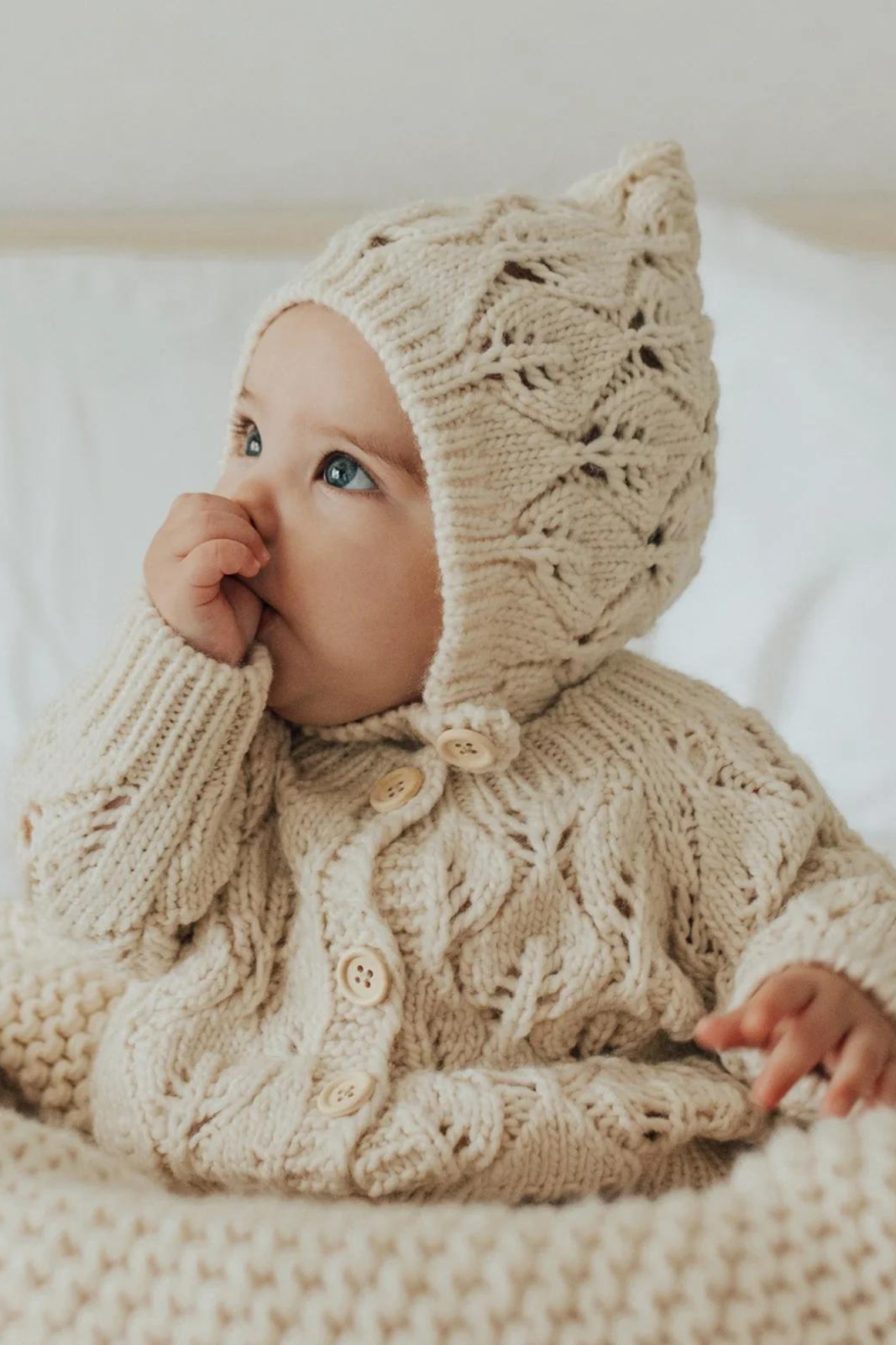 Baby in a beige knitted outfit and hat, sitting and sucking their thumb on a cozy blanket.