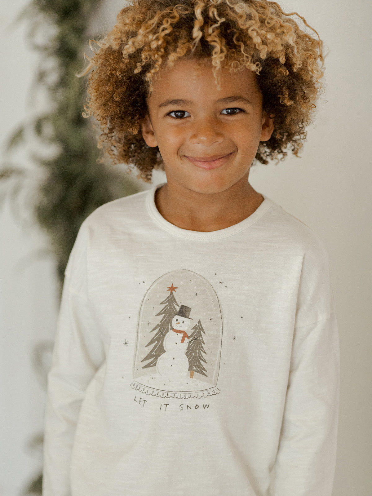 Smiling child wearing a festive long-sleeve shirt with a snowman design and "Let It Snow" text.
