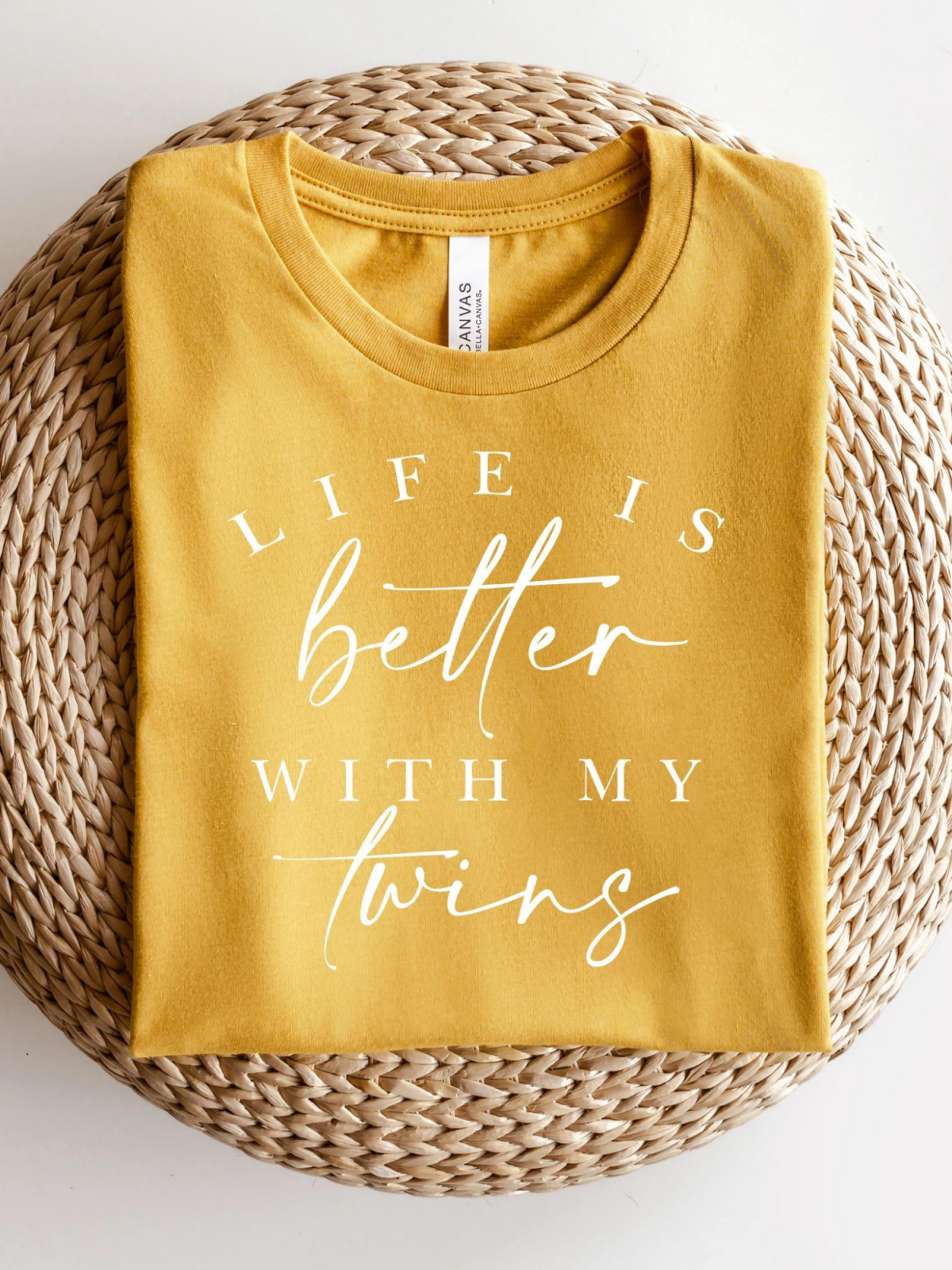 Yellow shirt folded on a woven placemat, with the text "Life is better with my twins" in white lettering.