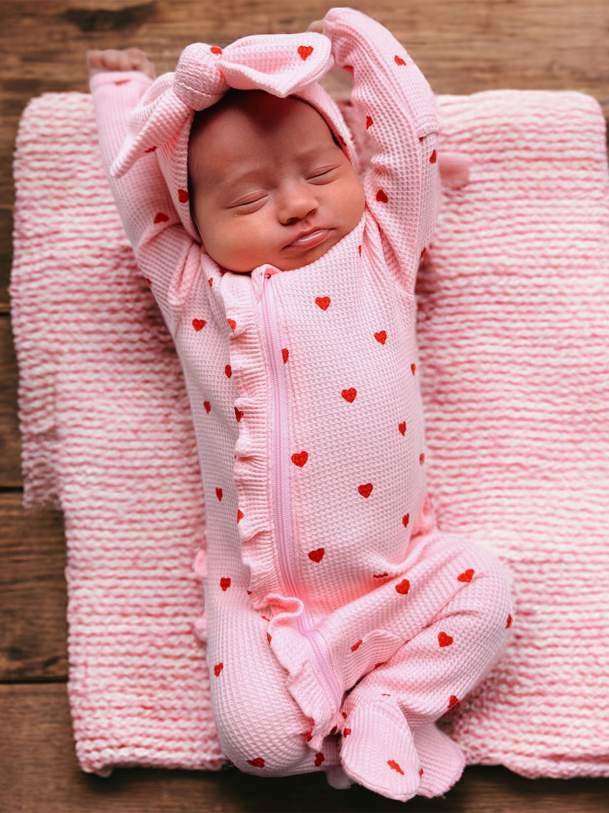 Newborn baby in pink heart-patterned outfit stretching on a pink blanket, eyes closed, looking peaceful and relaxed.