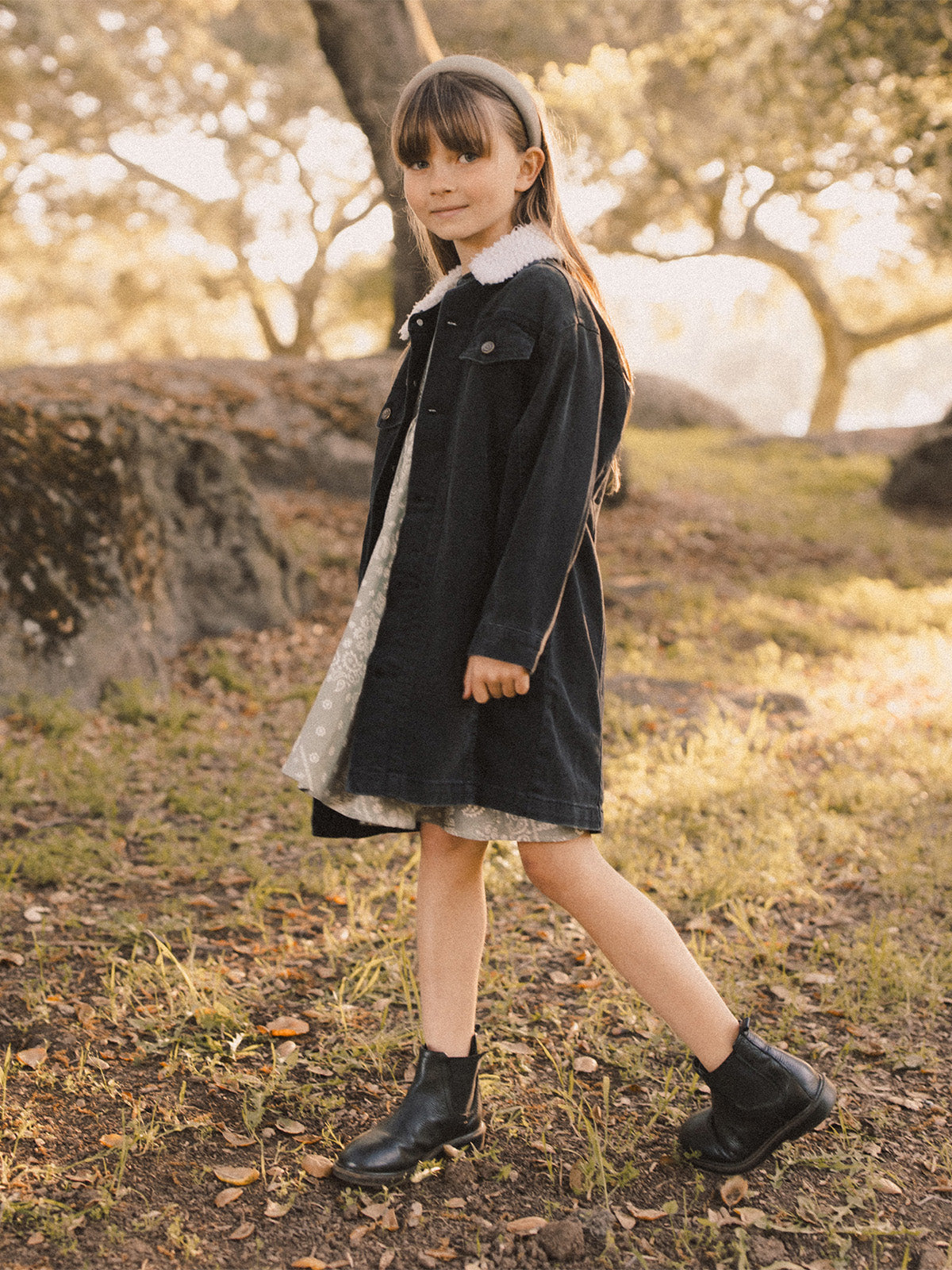 Girl wearing a black denim jacket and boots, standing outdoors in a grassy area with trees in the background.