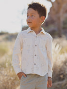 Smiling boy in a patterned shirt stands outdoors against a blurred natural background.