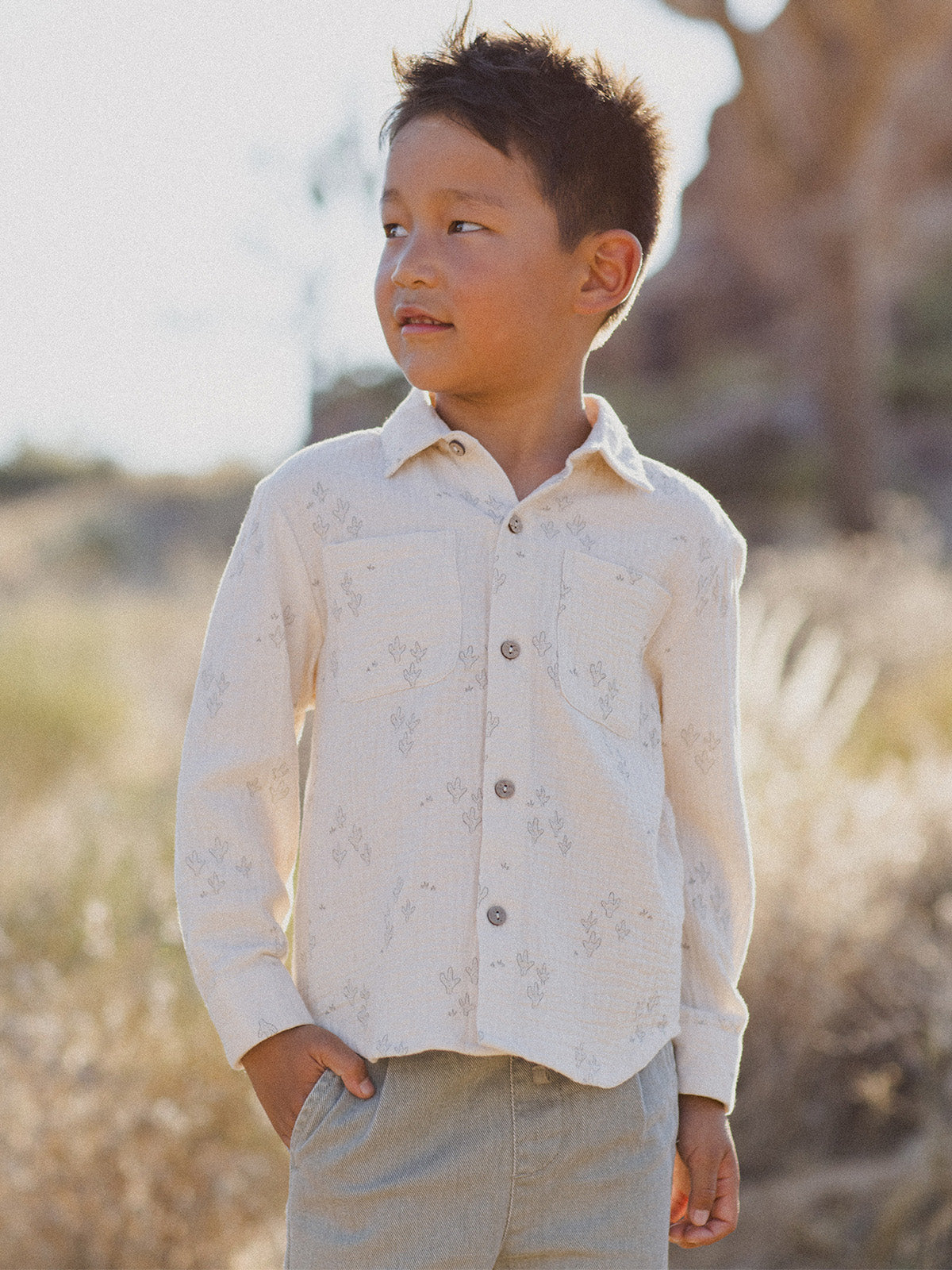 Smiling boy in a patterned shirt stands outdoors against a blurred natural background.