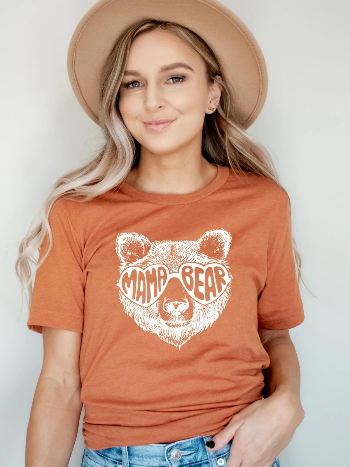 Woman smiling in a brown "Mama Bear" graphic tee, wearing a beige hat, against a plain background.