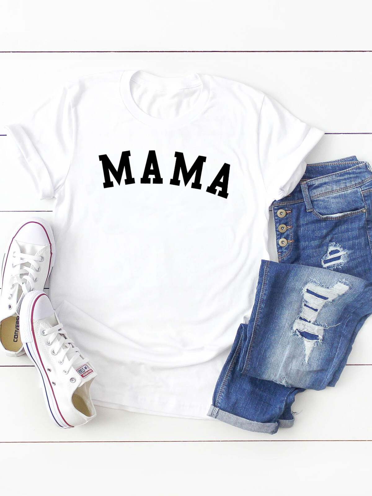 White t-shirt with "MAMA" text, paired with distressed blue jeans and white sneakers on a wooden surface.