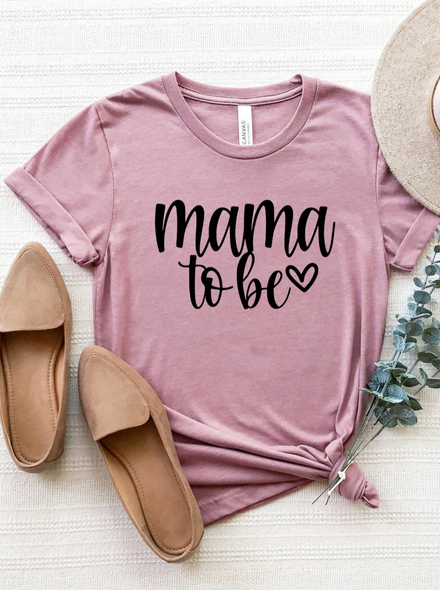 Pink t-shirt with "mama to be" text, paired with tan flats and greenery, styled on a textured background.