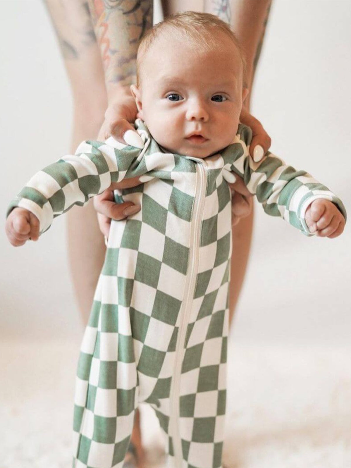 Baby in a green and white checkered onesie is held upright by an adult, showcasing a playful, cozy look.