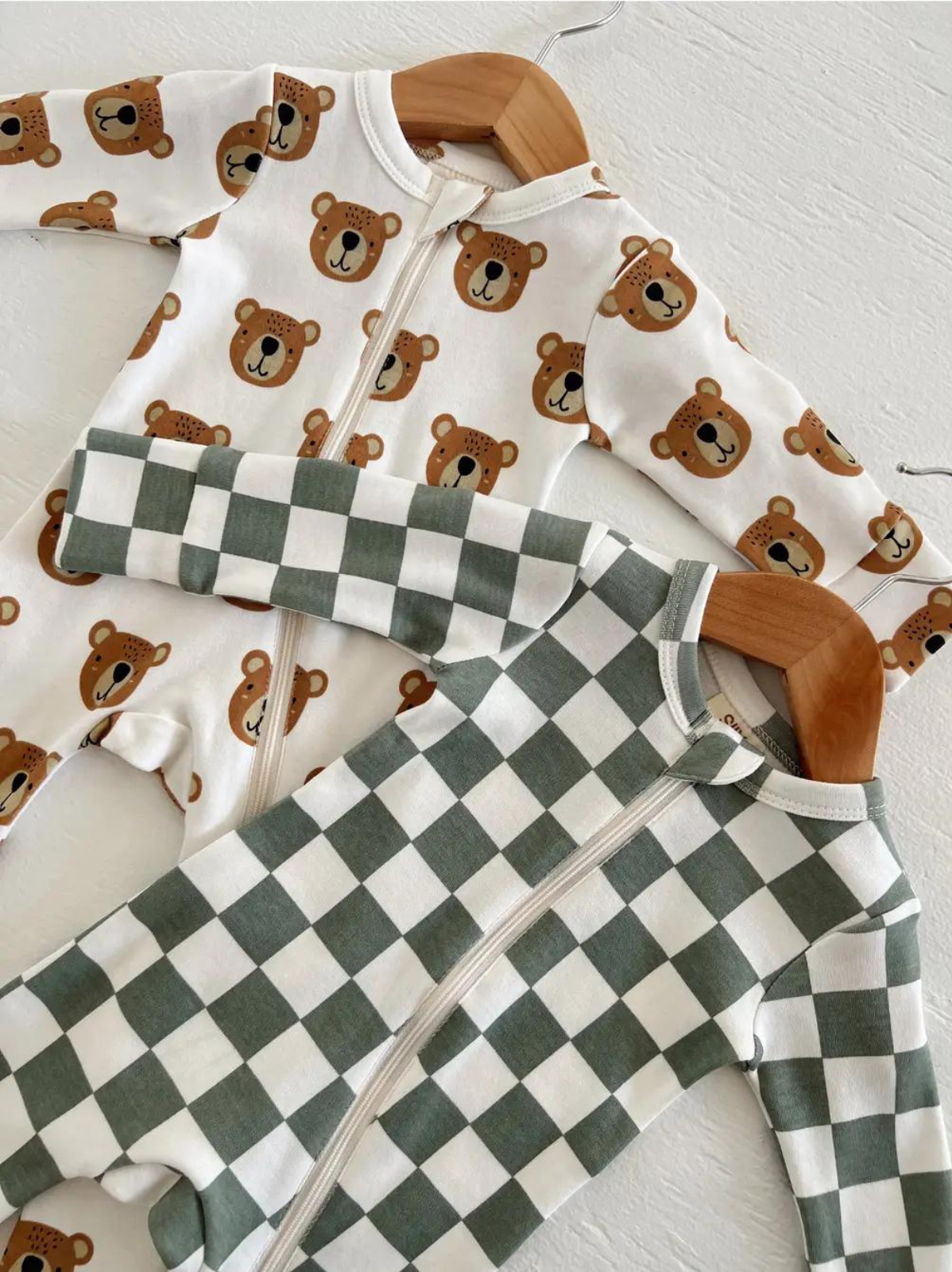 Two baby onesies, one with bear print and the other in green checkered pattern, hanging on wooden hangers.