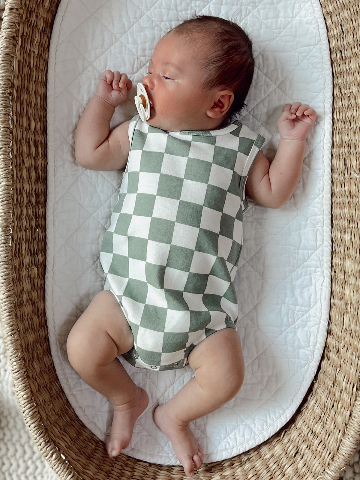 Baby wearing a green and white checkered onesie, lying in a woven basket with a pacifier.