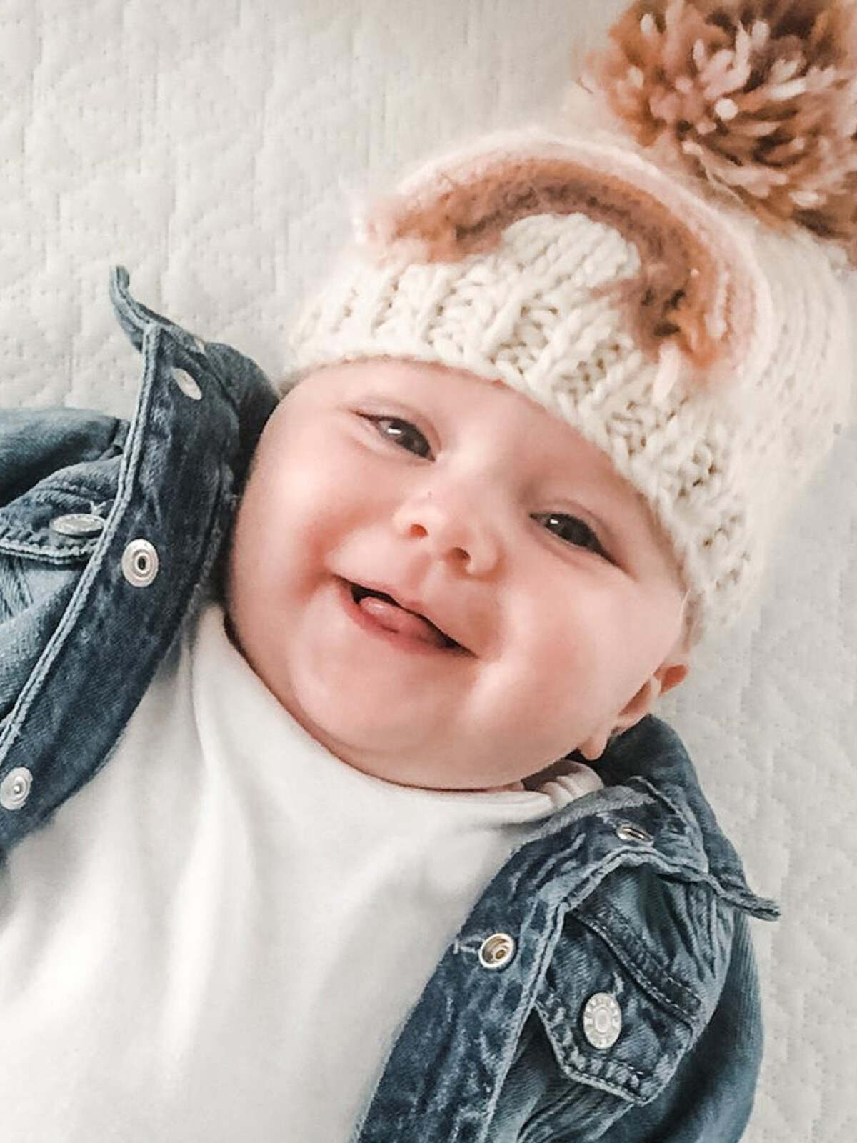 Smiling baby in a knitted hat and denim jacket, lying on a soft blanket.