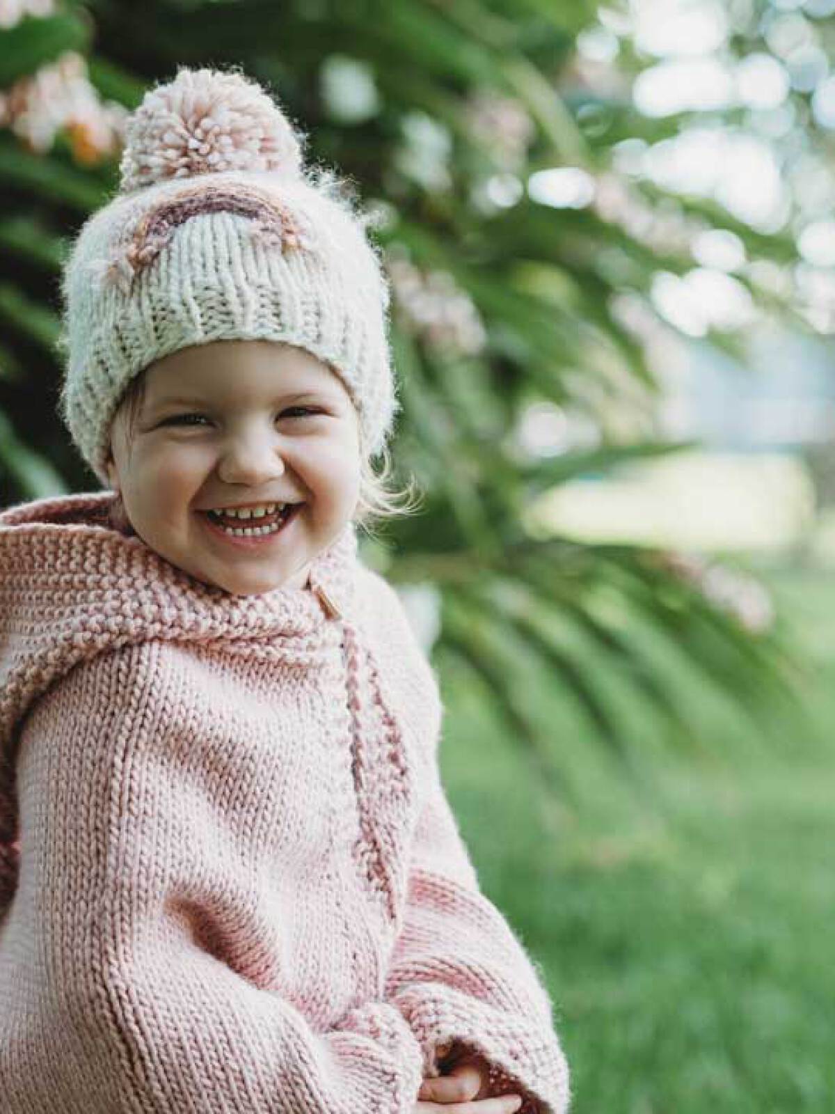 Smiling child in a cozy pink sweater and knitted hat, surrounded by green foliage.