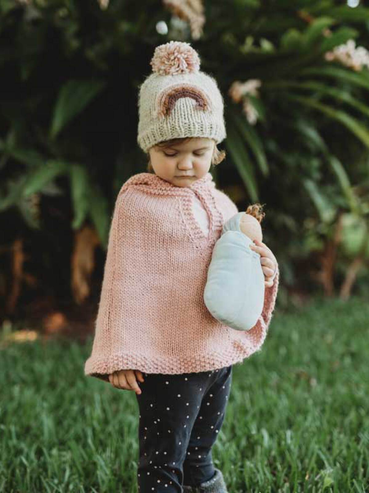 Child wearing a pink knitted poncho and hat, holding a doll, standing on green grass with foliage in the background.