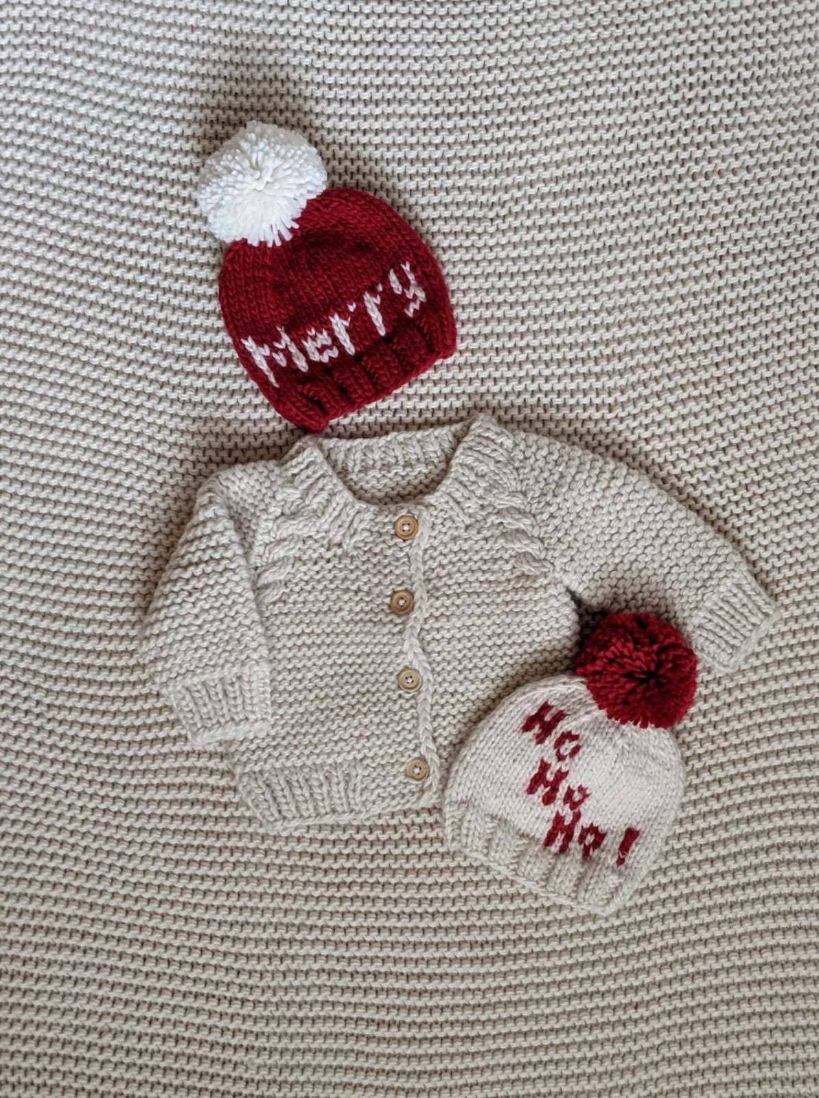 Baby cream cardigan with two holiday-themed knitted hats, one red with "Merry" and one white with "Ho Ho Ho!"