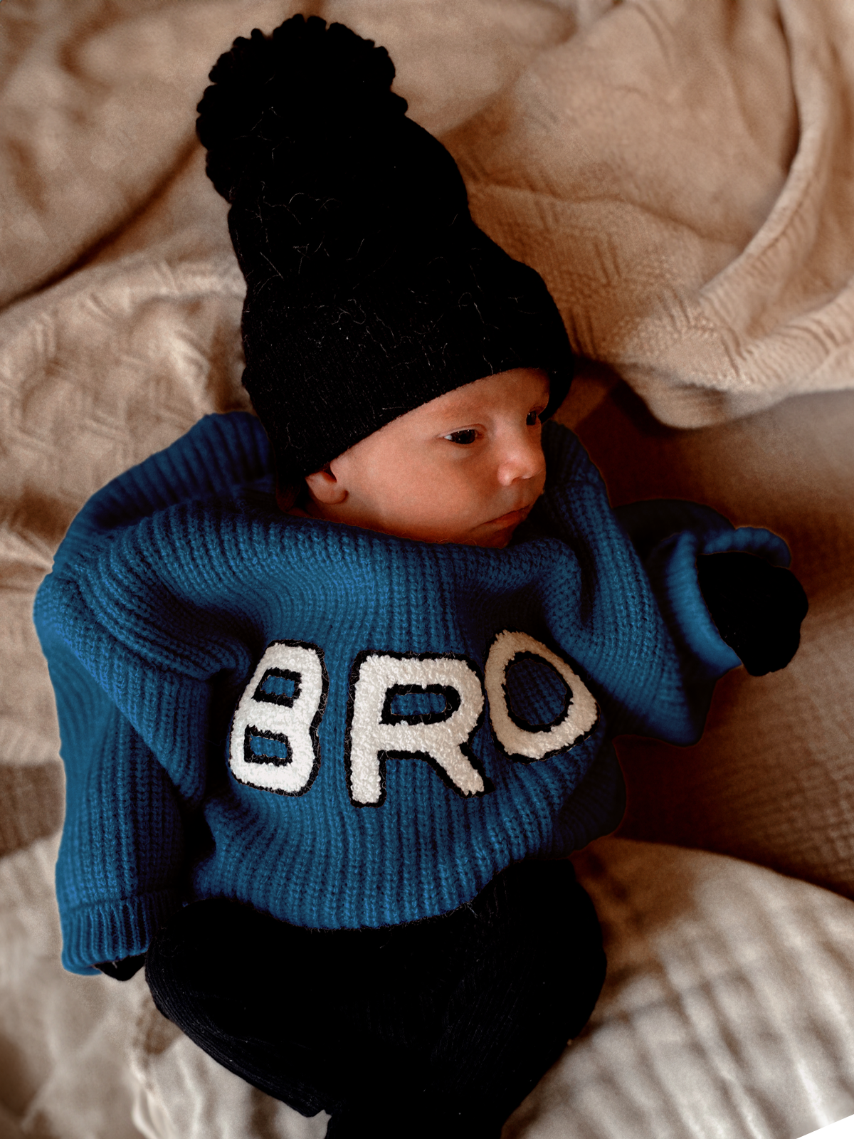 A baby in a blue sweater with "BRO" text, wearing a black beanie, rests on a blanket.