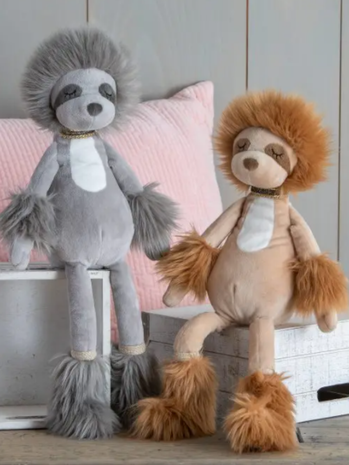 Two plush animal toys, a gray sloth and a tan lion, seated on decorative boxes with a pink pillow in the background.