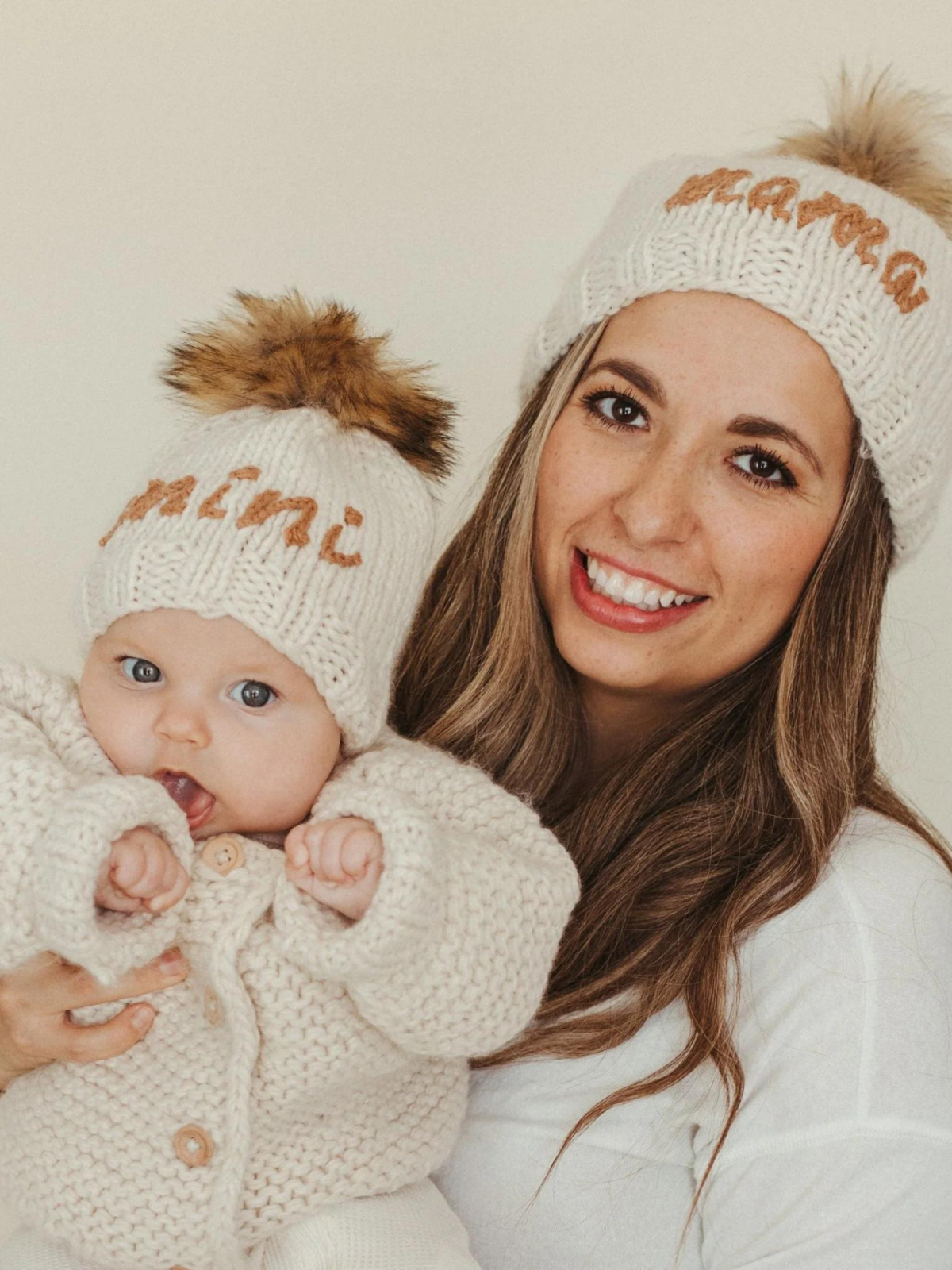 Mother and baby in matching knit hats, smiling together against a neutral background.