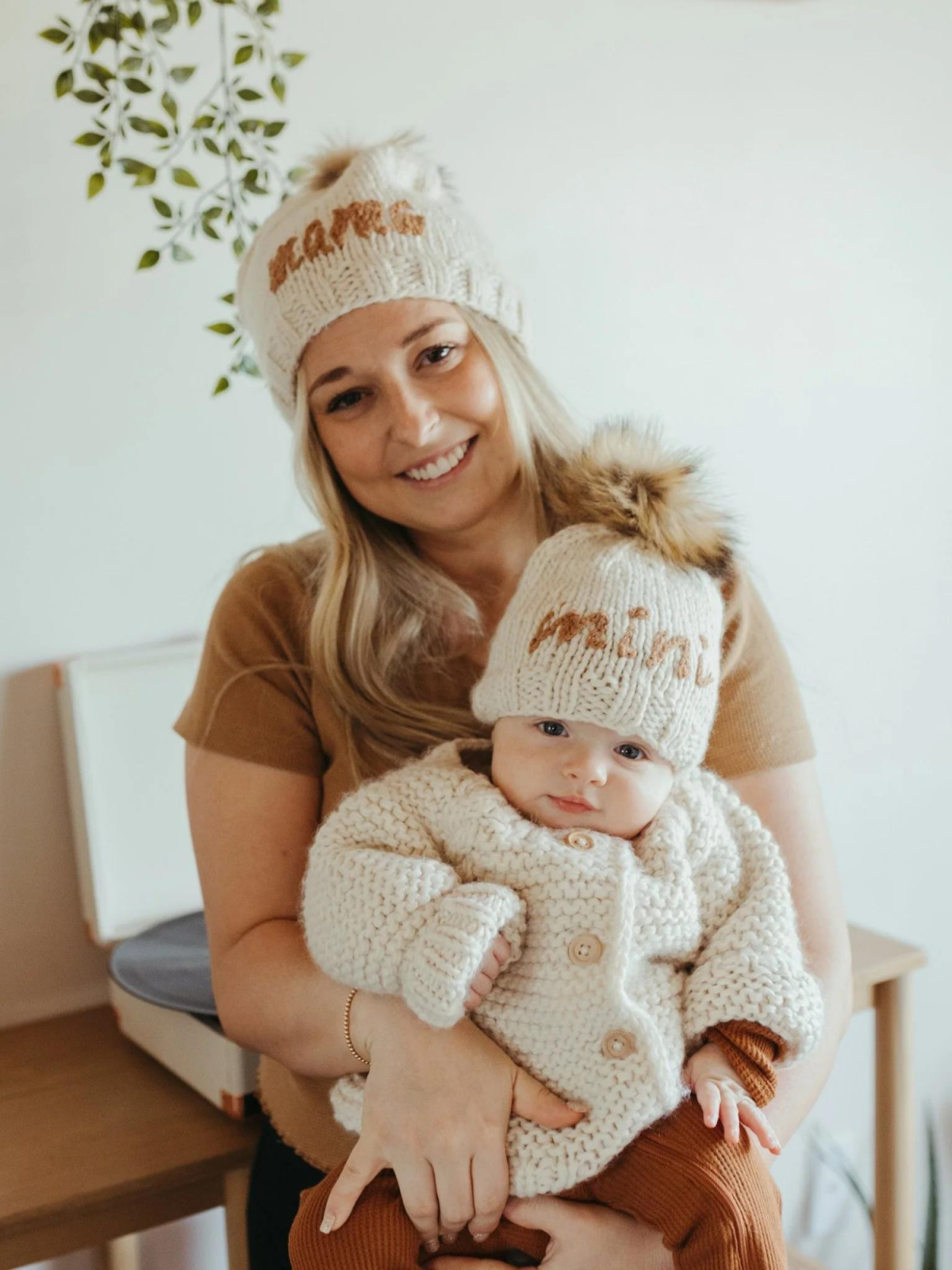 Mom holding a baby, both in cozy knit hats, smiling in a softly lit room with greenery in the background.