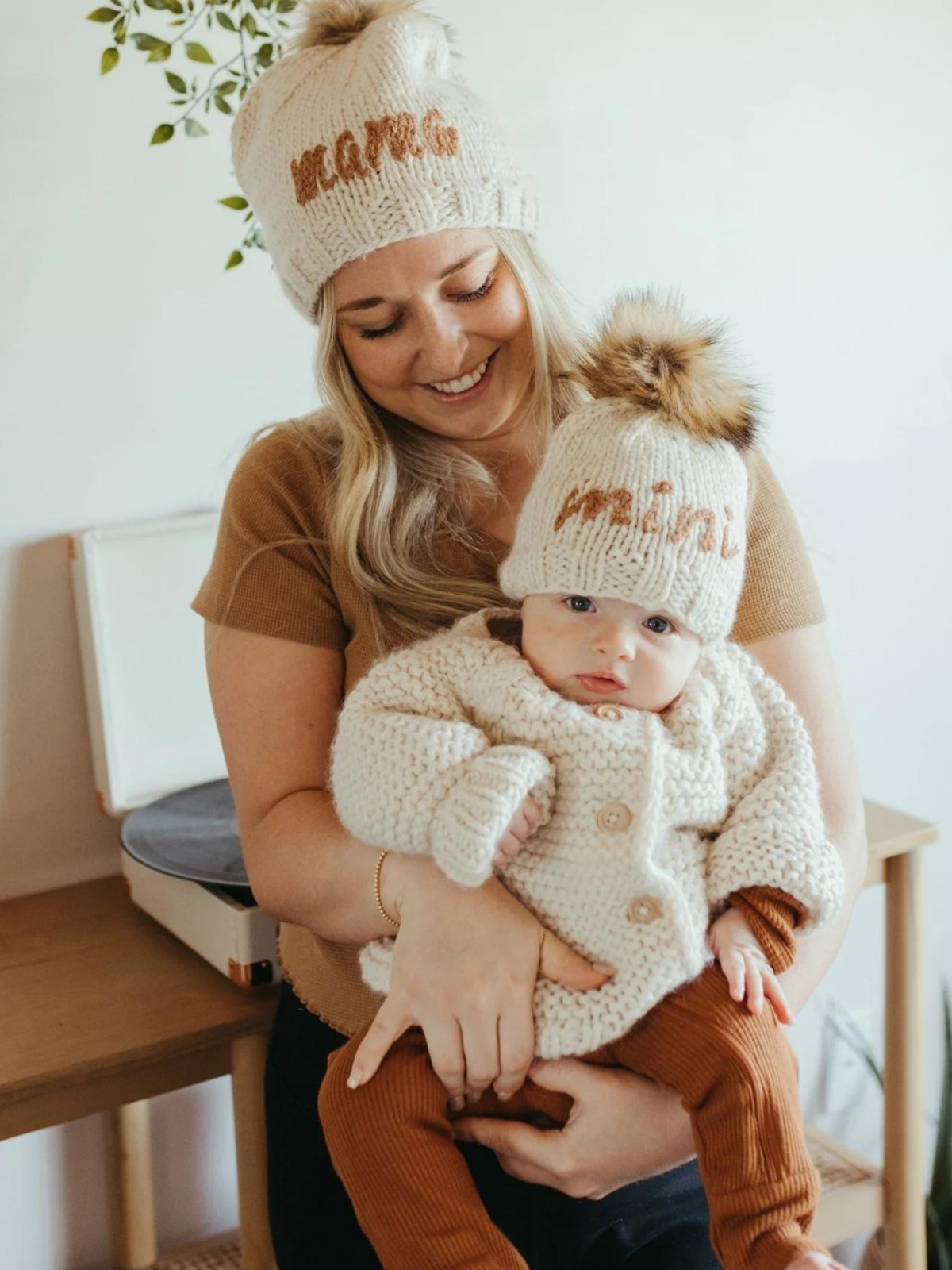 Mother and baby wearing matching knitted hats, with the words "mom" and "mini," smiling at each other indoors.
