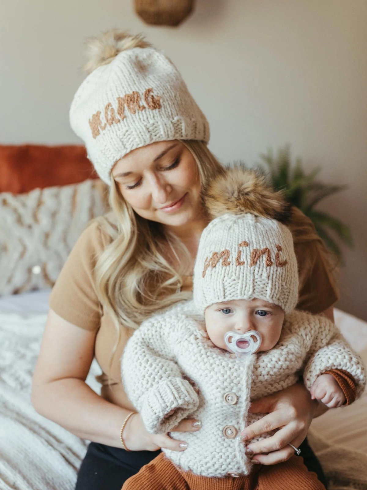 Mom and baby both wear matching knitted hats, with "MAMA" and "MINI" text, cozy setting with warm colors.