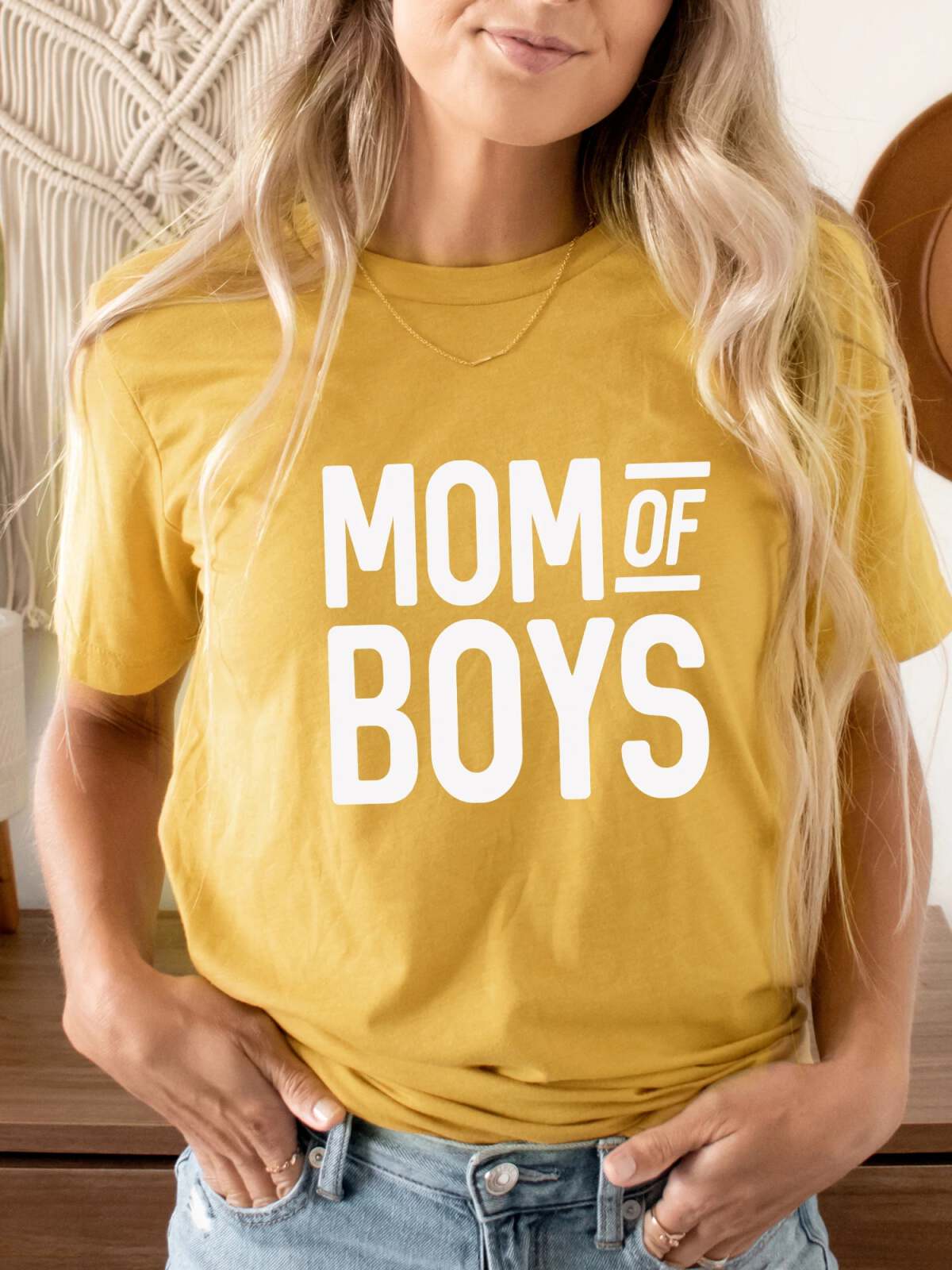 Woman wearing a yellow t-shirt with "MOM OF BOYS" text, standing in a cozy room.