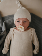 Newborn baby with pacifier wearing a beige outfit and hat, resting on a soft gray blanket.