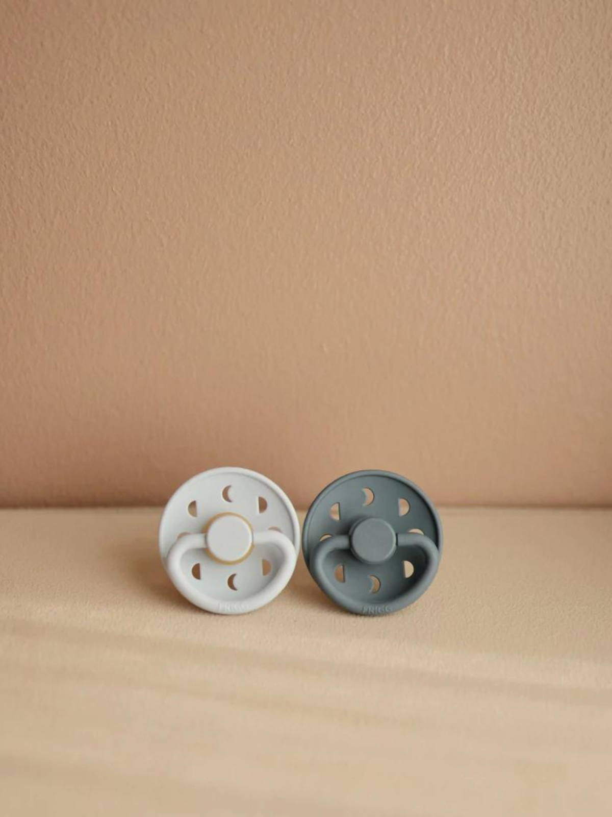 Two pacifiers, one white with gold accents and one gray, placed on a beige surface against a soft background.