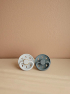 Two pacifiers, one white with gold accents and one gray, placed on a beige surface against a soft background.