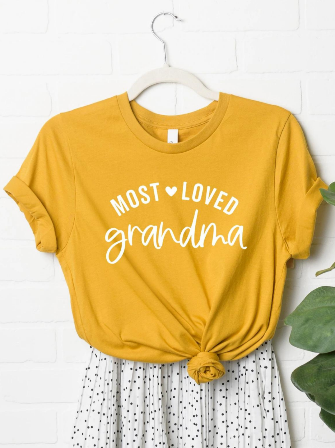 Yellow t-shirt with "Most Loved Grandma" text, knotted at the waist, hanging on a white wall with plants.