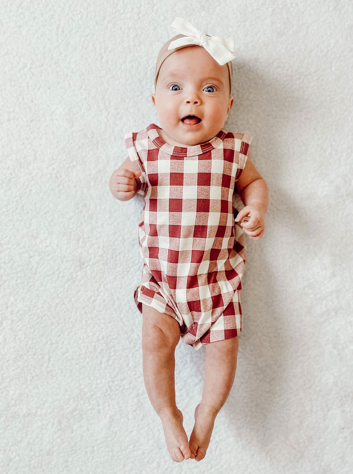 Baby lying on a soft surface, smiling, wearing a red and white checked romper and a matching headband.