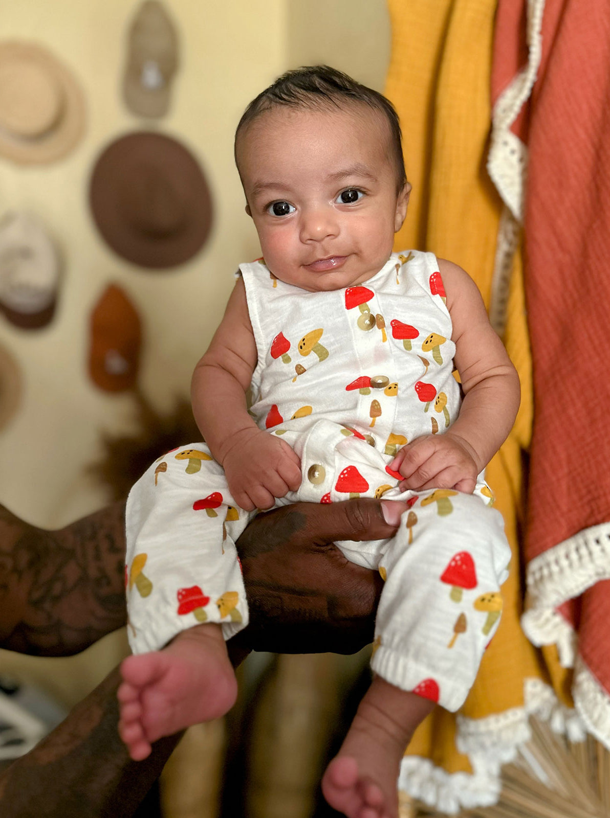 Smiling baby in a playful outfit with mushrooms and strawberries, held against a colorful background.