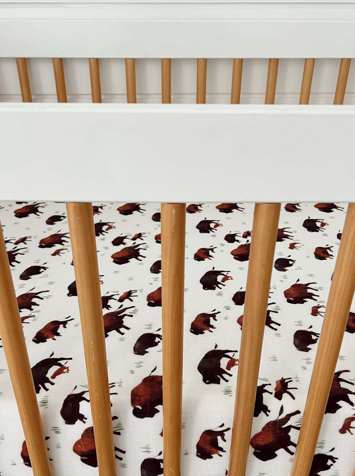 White crib with wooden slats and a patterned sheet featuring bison prints on a light background.