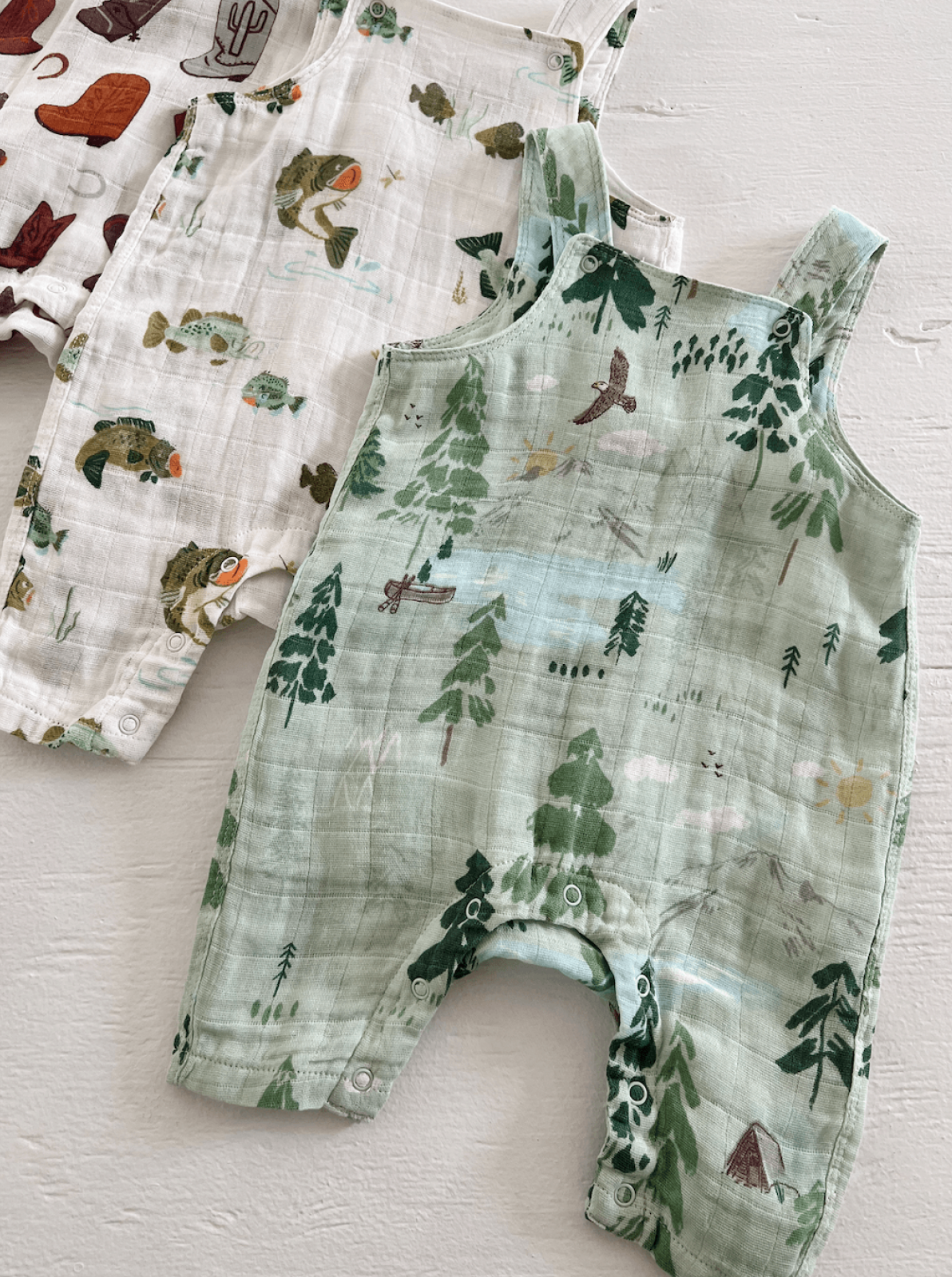 Three colorful baby rompers with nature-themed prints: fish, trees, and camping scenes, on a light background.