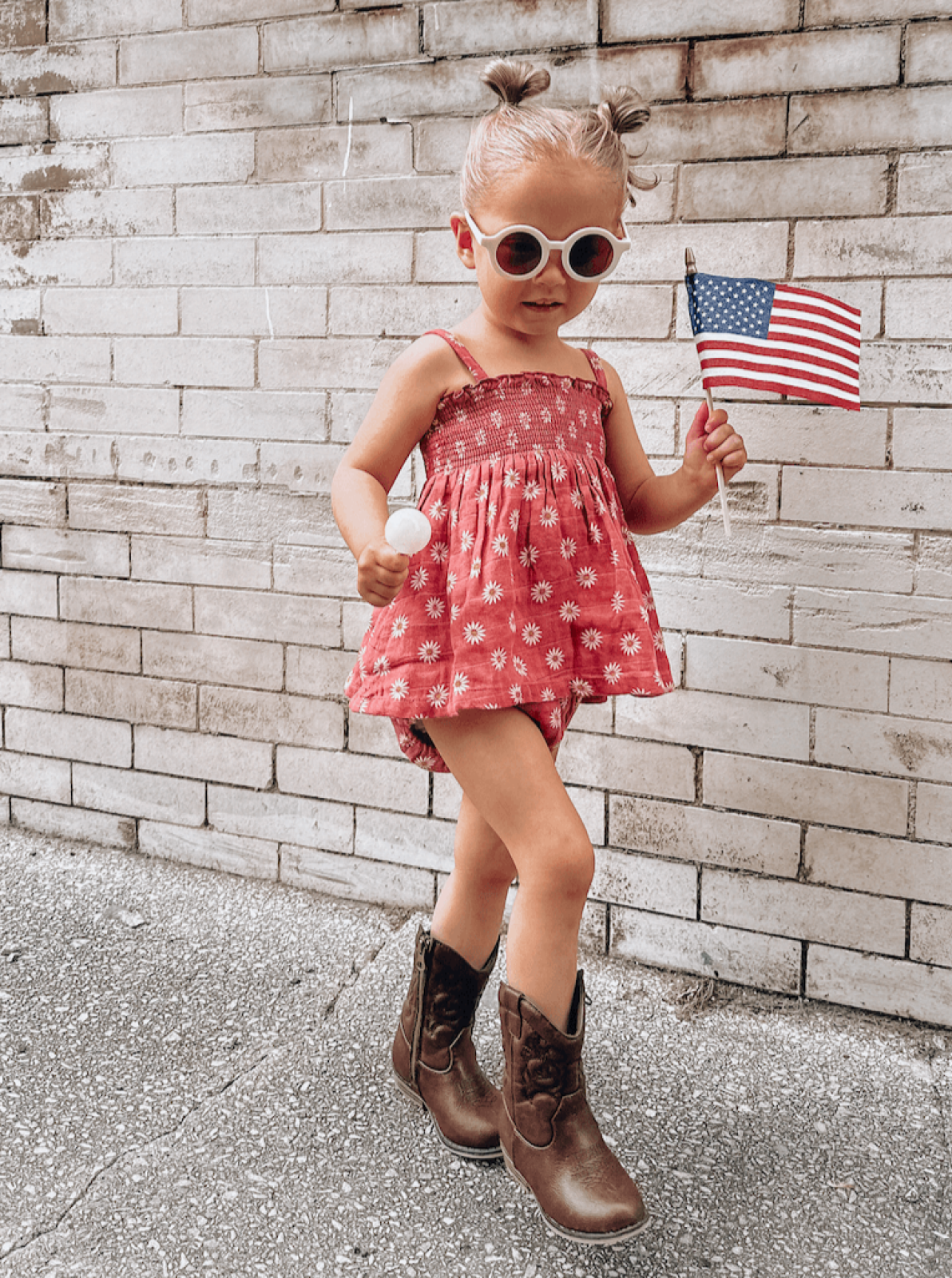 Young girl in a floral dress and sunglasses holding an American flag, smiling against a brick wall background.