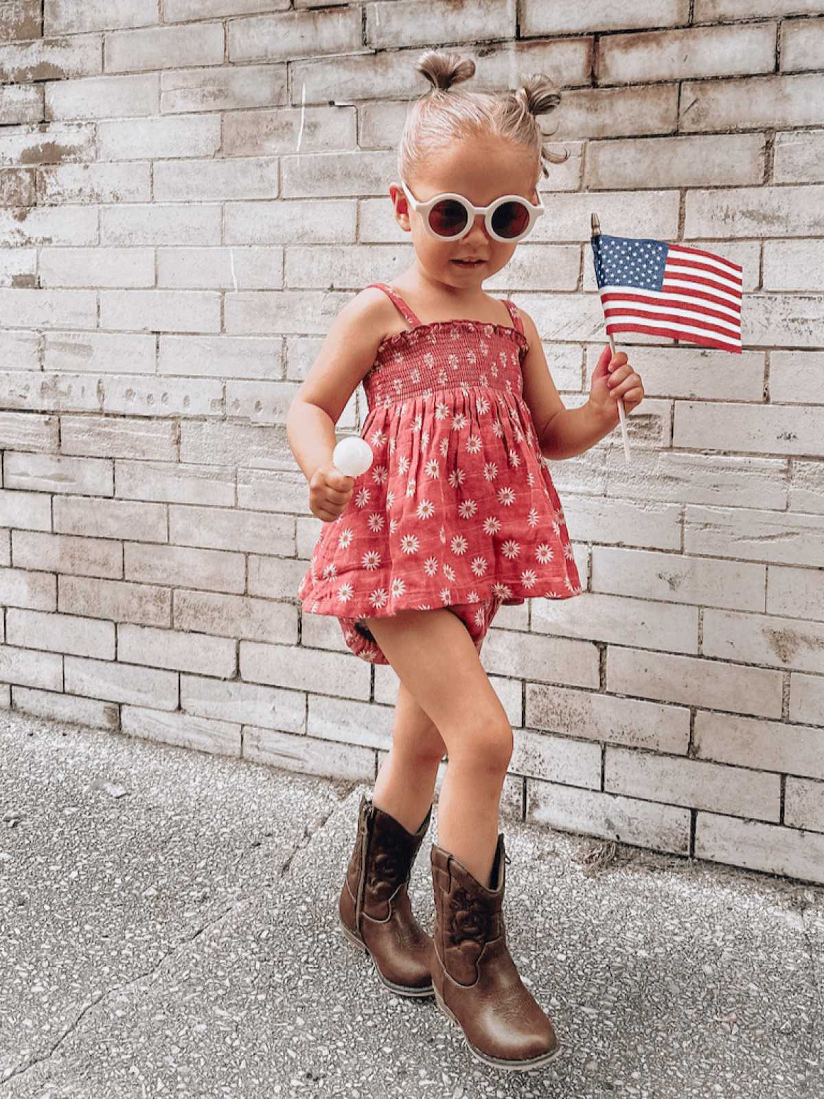 Young girl in a floral dress and sunglasses holding an American flag, smiling against a brick wall background.