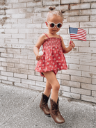 Young girl in a floral dress and sunglasses holding an American flag, smiling against a brick wall background.