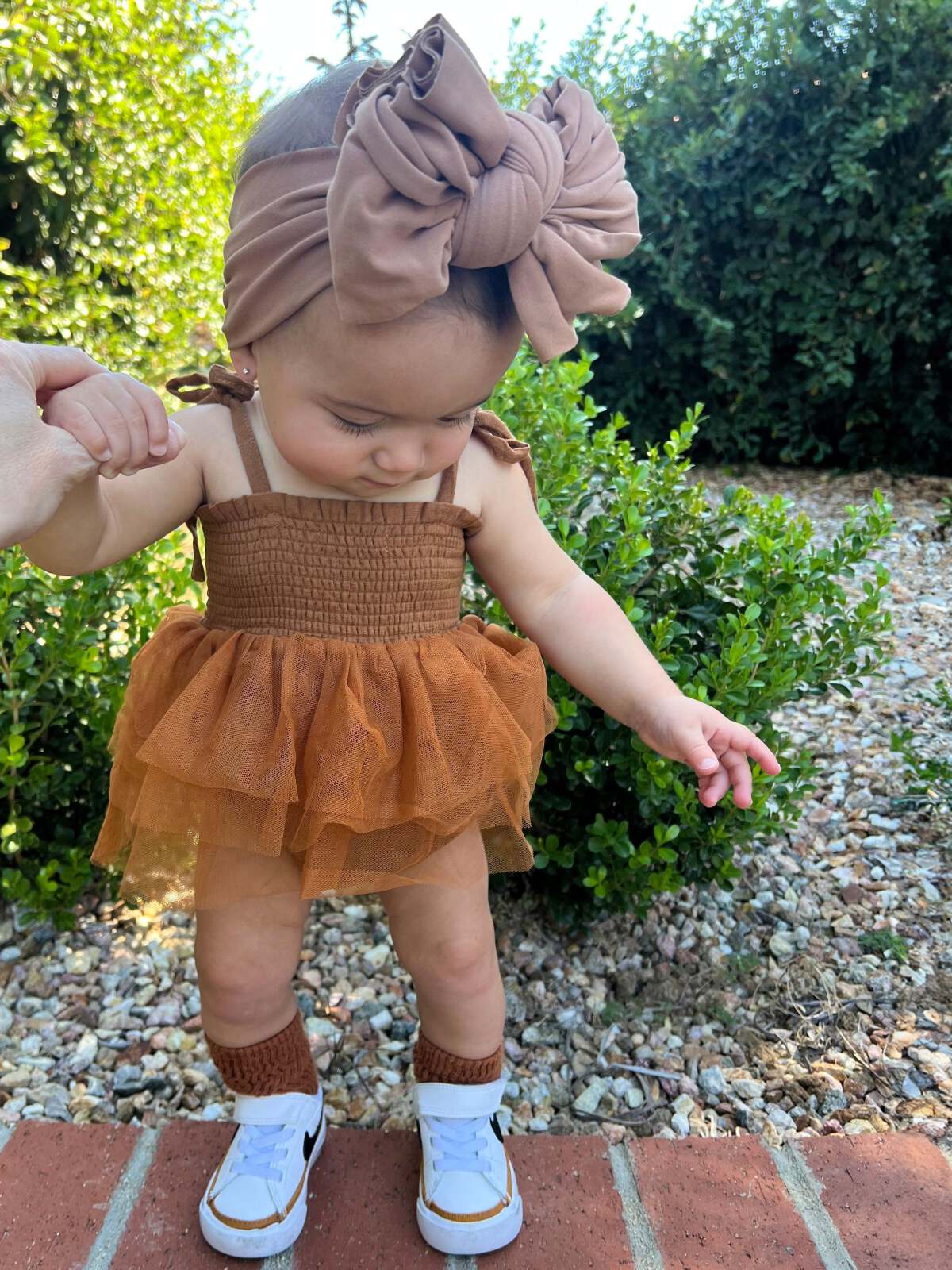 Baby girl in a brown dress and headband, standing outside with greenery in the background.
