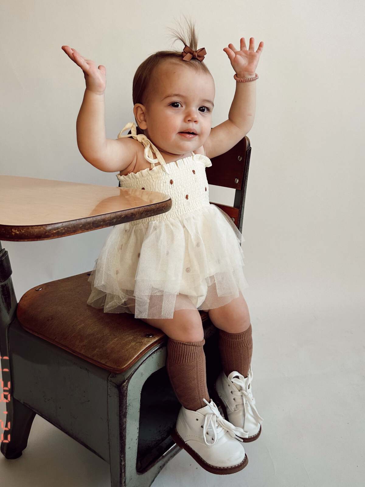 Toddler in a cream dress and knee-high socks sitting on a chair, raising hands with a joyful expression.