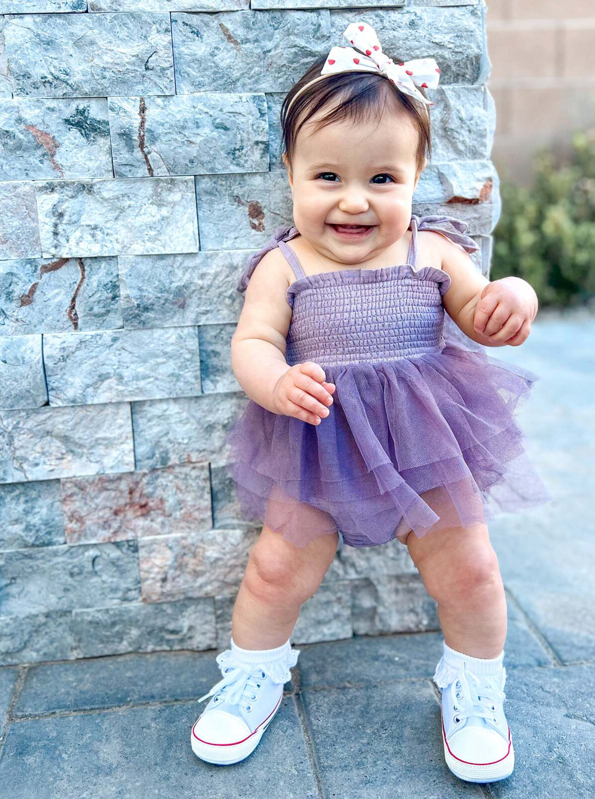 Smiling baby wearing a purple dress and white sneakers, standing in front of a stone wall.