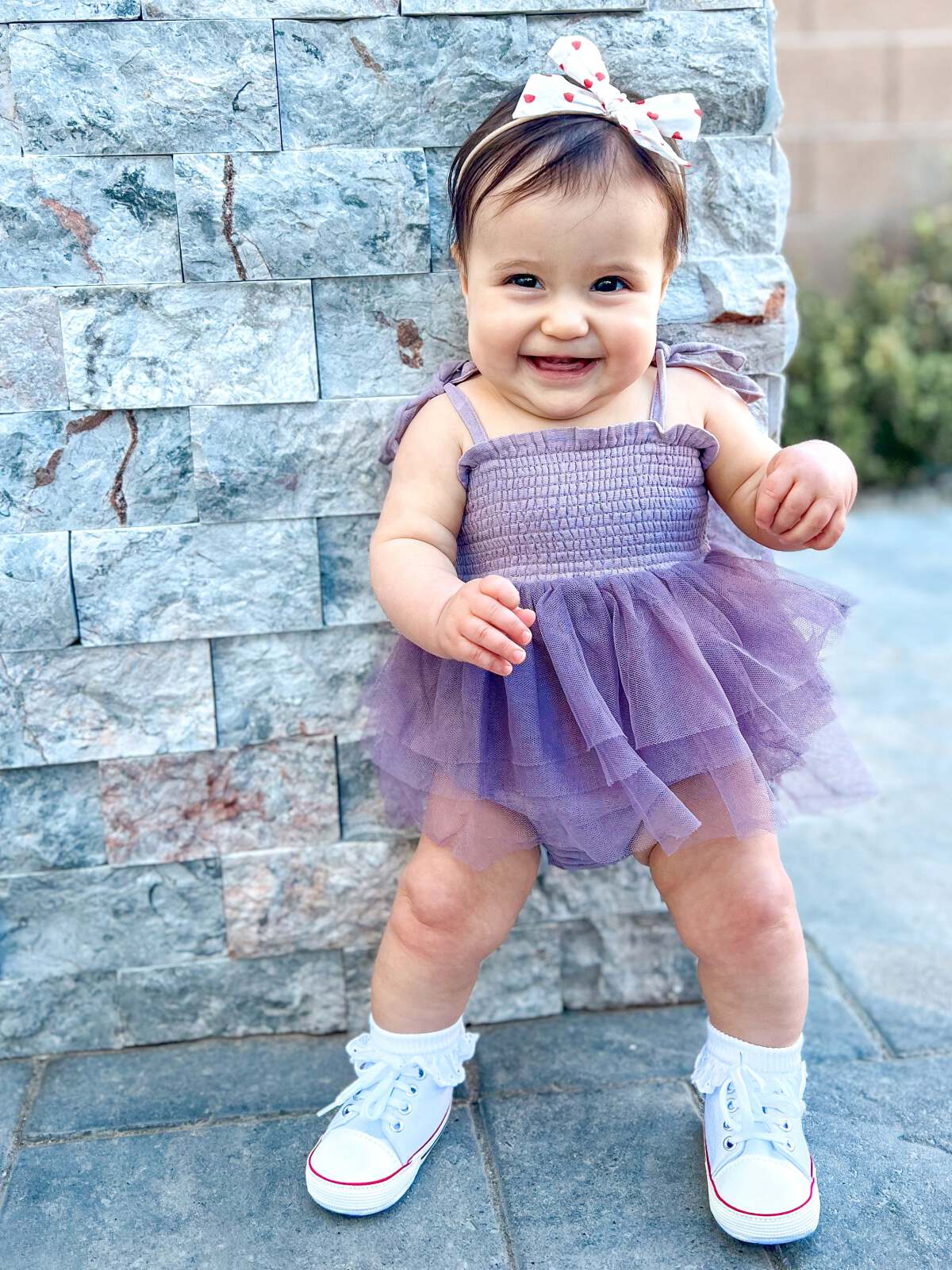 Smiling baby wearing a purple dress and white sneakers, standing in front of a stone wall.