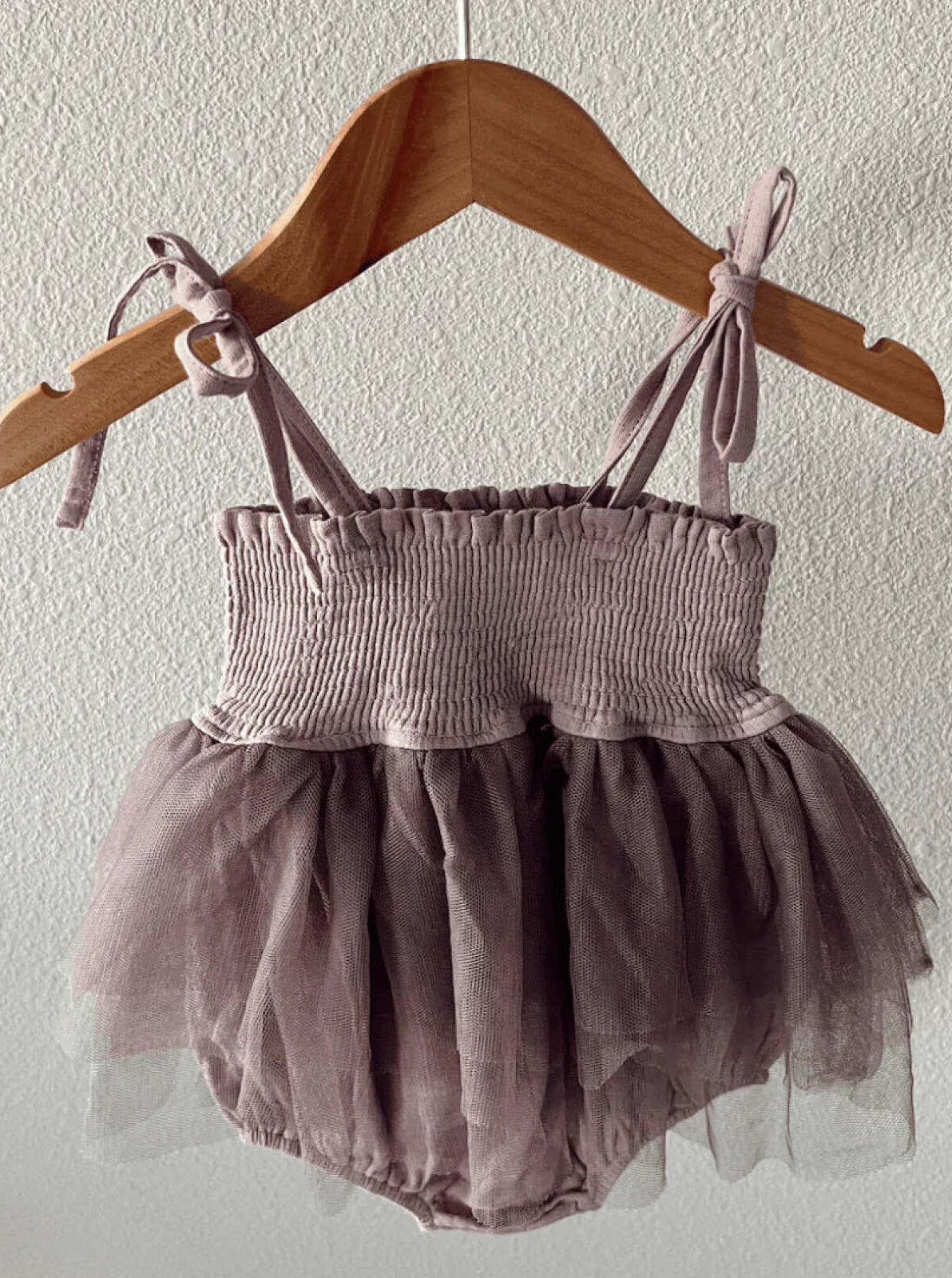 Dusty purple baby romper with a smocked bodice and tulle skirt, hanging on a wooden hanger against a wall.