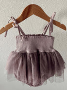 Dusty purple baby romper with a smocked bodice and tulle skirt, hanging on a wooden hanger against a wall.