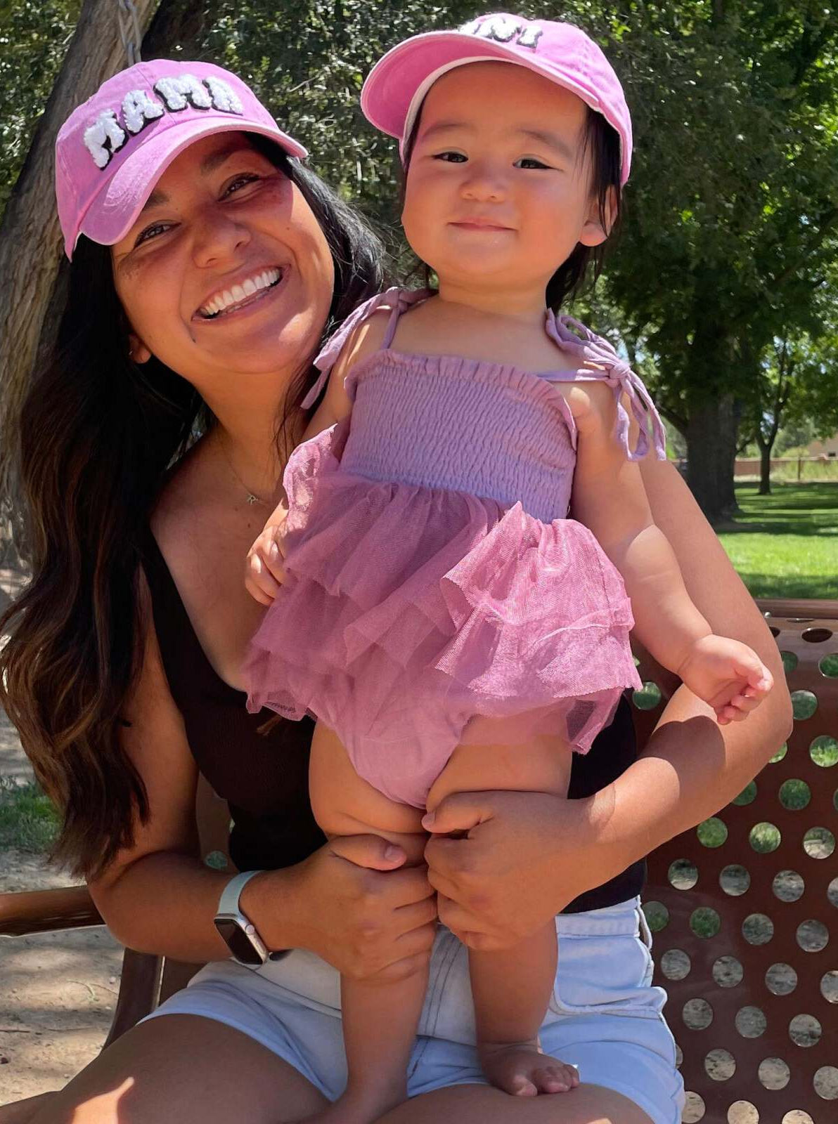 Smiling woman in a pink hat holds a happy baby in a frilled pink outfit, outdoors in a park.