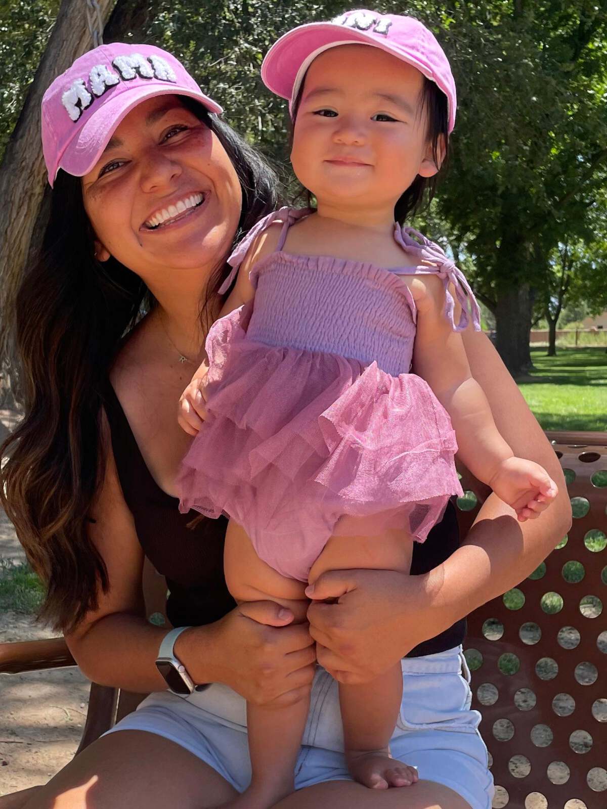 Smiling woman in a pink hat holds a happy baby in a frilled pink outfit, outdoors in a park.