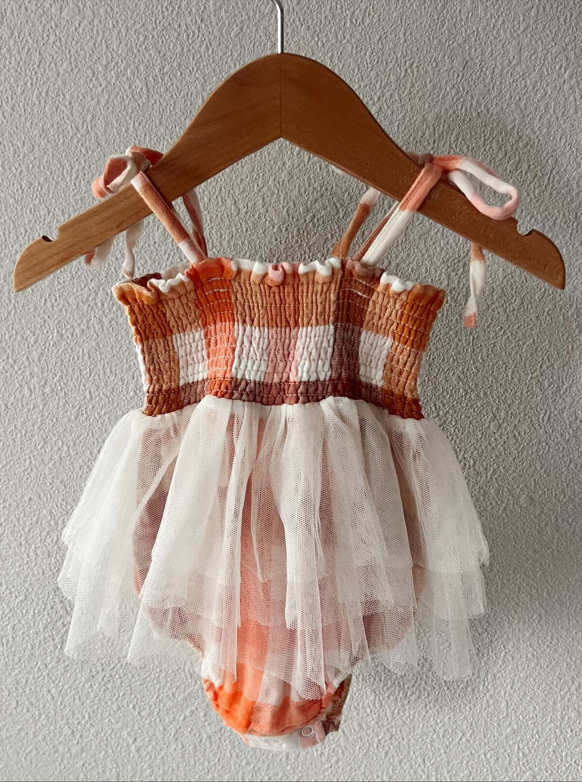 Peach and white checked toddler swimsuit with a tulle skirt, hanging on a wooden hanger against a textured wall.