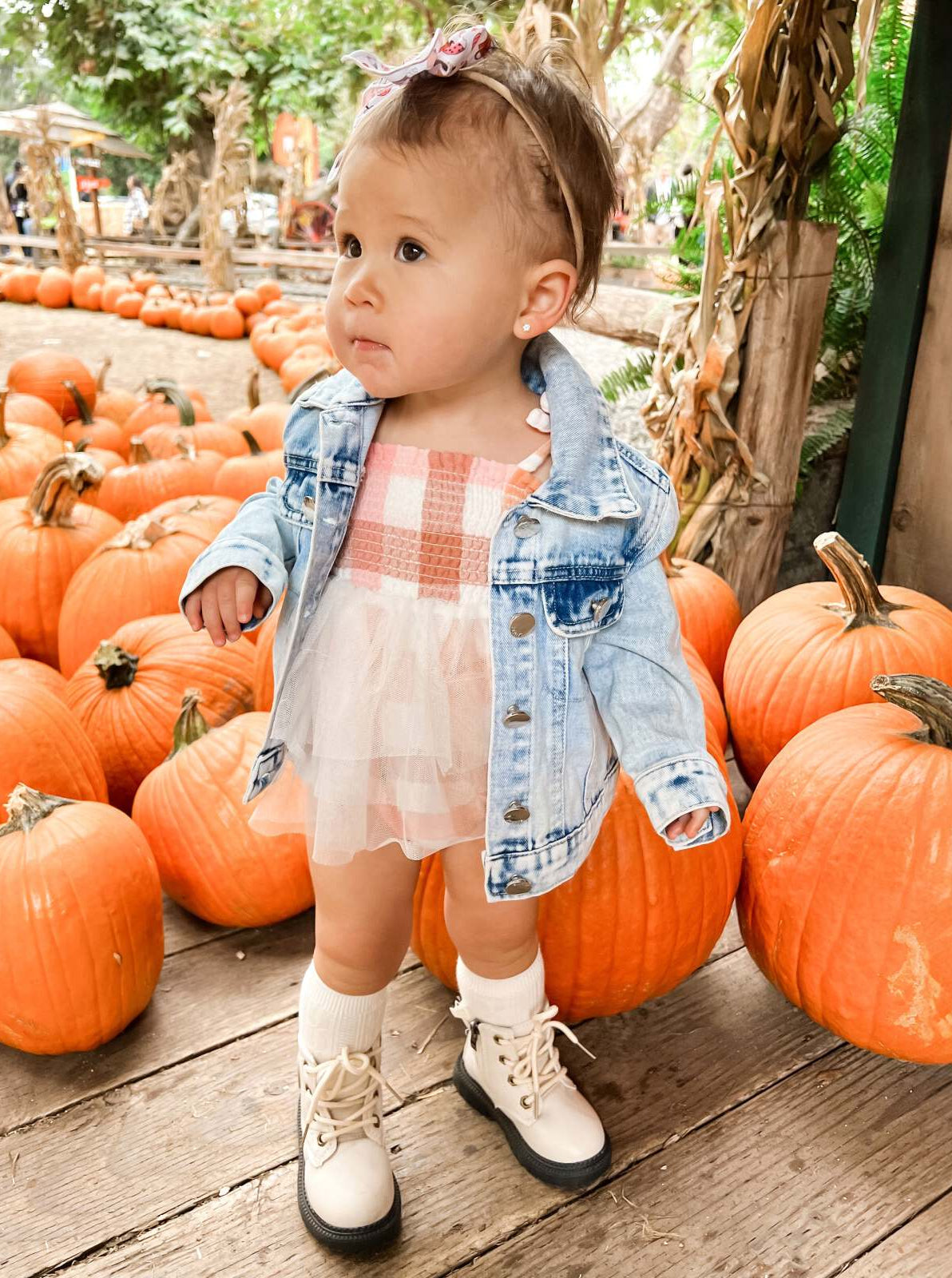 Toddler in a denim jacket stands by pumpkins, wearing white boots and a pink dress, with a bow in her hair.