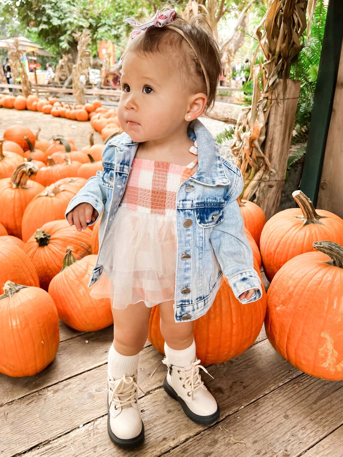 Toddler in a denim jacket stands by pumpkins, wearing white boots and a pink dress, with a bow in her hair.