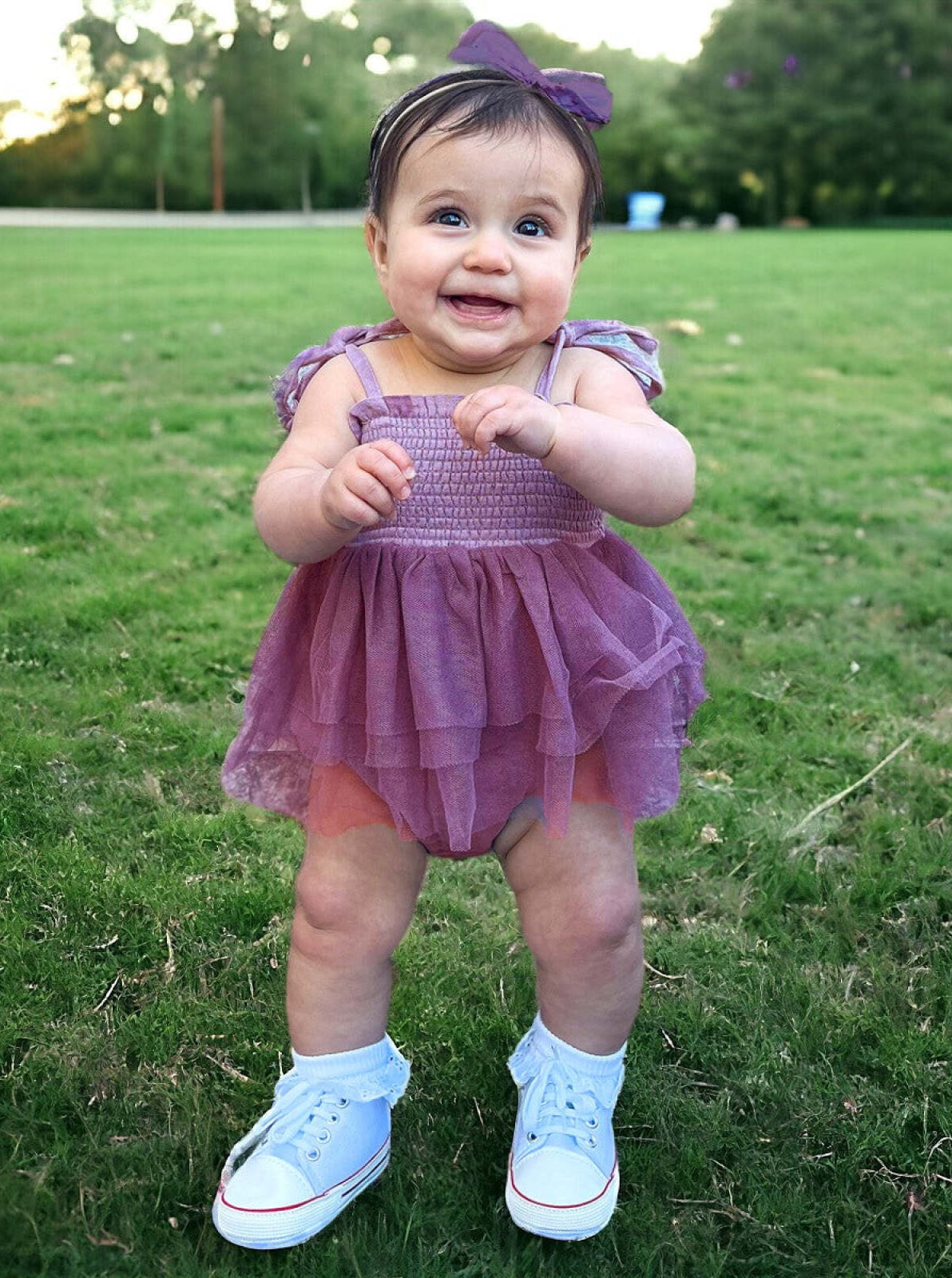 Smiling baby girl in a purple dress and white shoes standing on grass, looking joyful and playful.