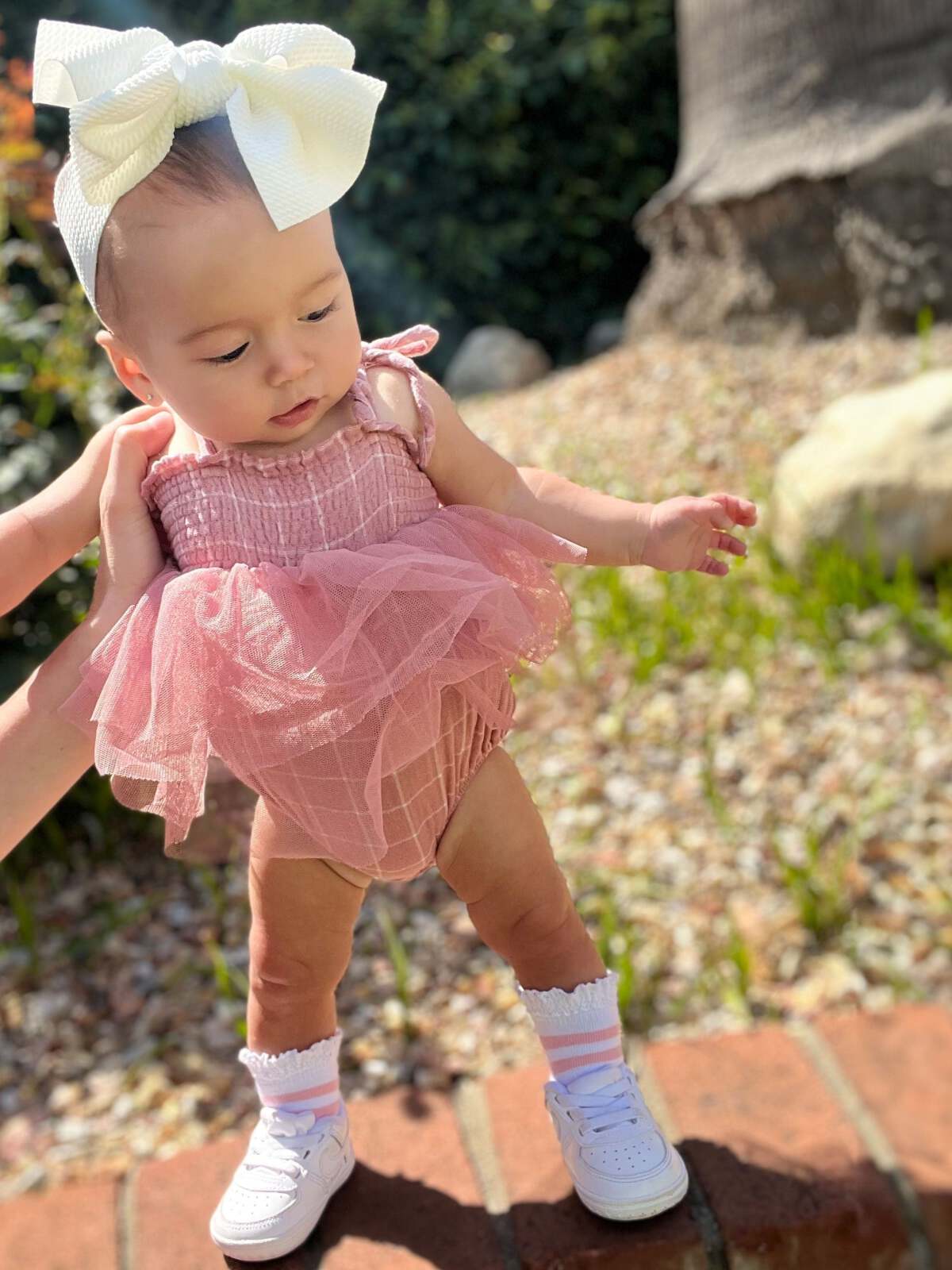 Baby in a pink tutu outfit with a large white bow, standing outdoors on a sunny day.