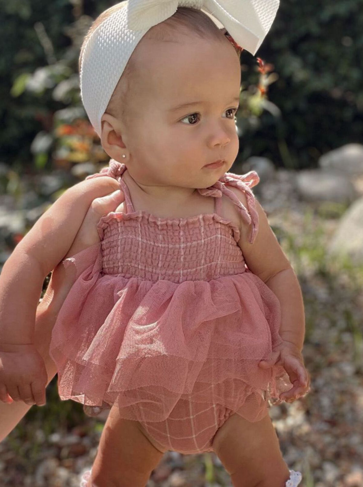 Toddler in a pink outfit with a bow headband, standing outdoors with greenery in the background.